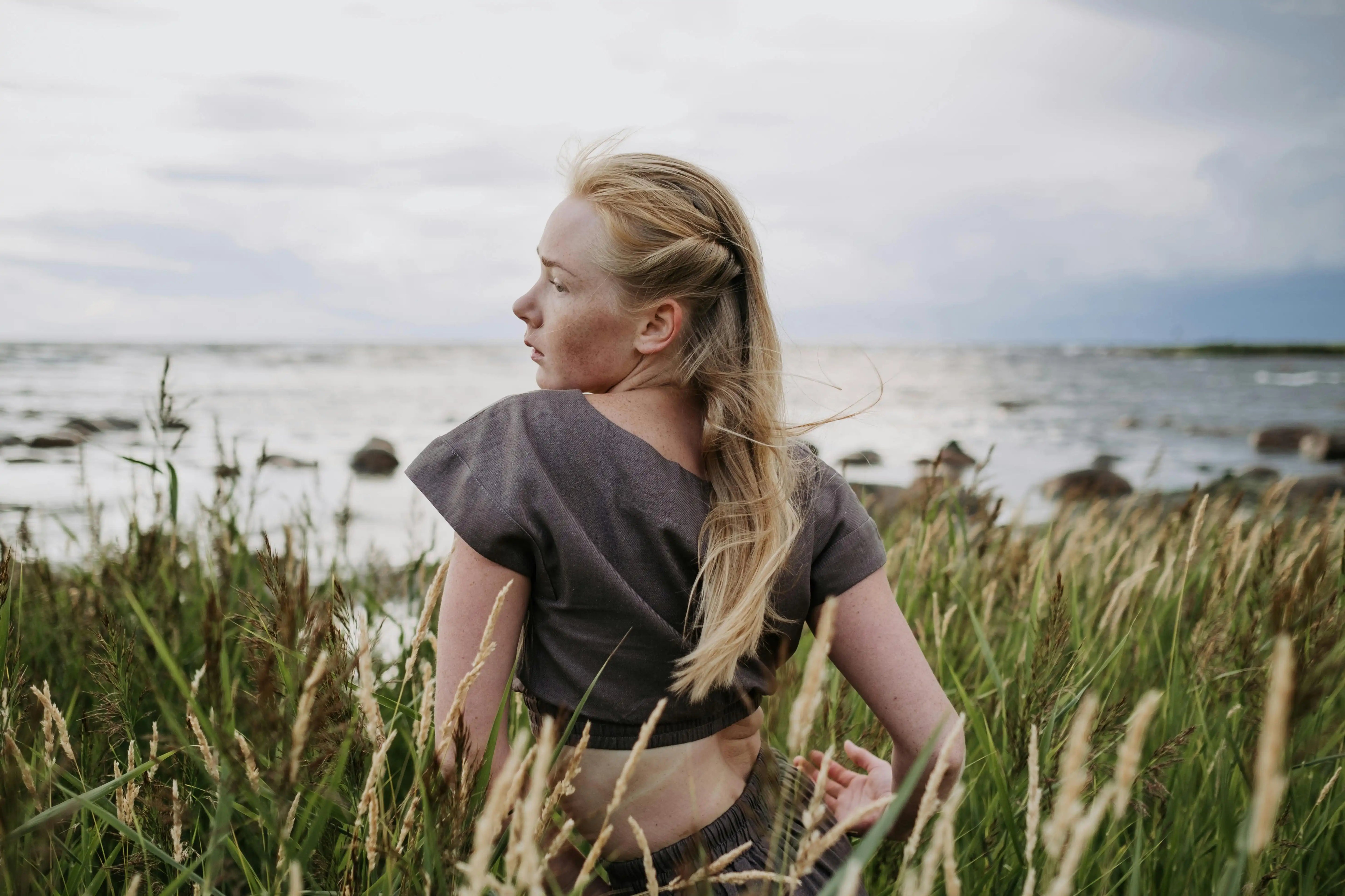 Frau mit langen blonden Haaren, die unter bewölktem Himmel auf einer Wiese an einer felsigen Küste sitzt.