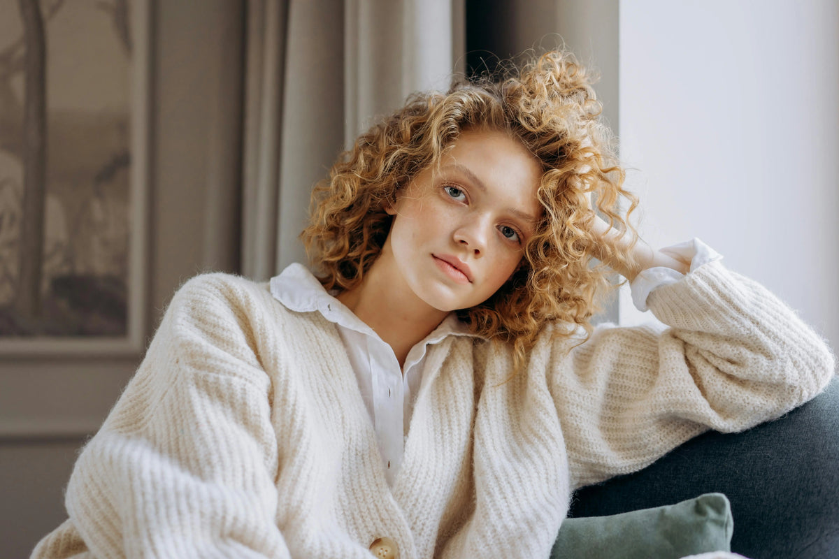 Junge Frau mit lockigem Haar in einem weißen Pullover, die auf einer Couch neben einem Fenster sitzt.