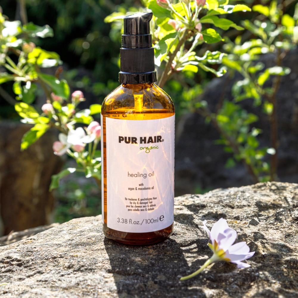 PUR HAIR Organic Healing Oil in brauner Glasflasche auf Steinoberfläche vor Blütenbusch