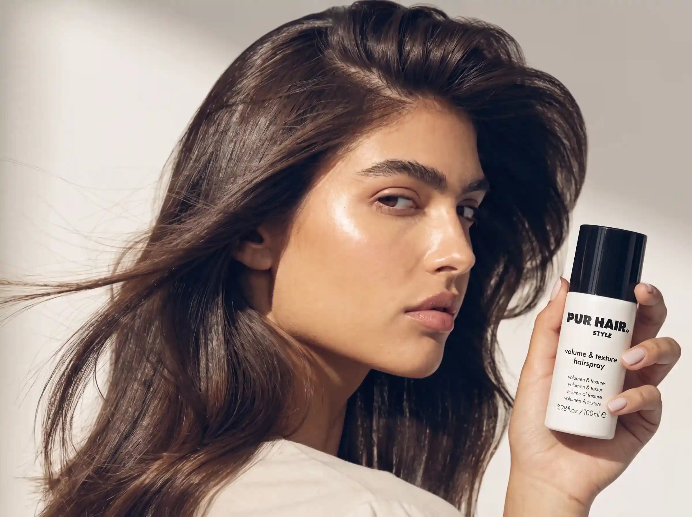 Model mit voluminösem glänzendem braunem Haar hält PUR HAIR Volume & Texture Haarspray neben das Gesicht.