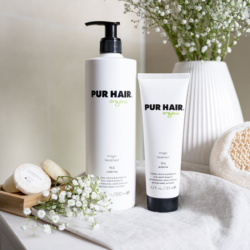 Weiße Flaschen PUR HAIR Organic Magic Treatment 1000 ml und 125 ml, auf grauem Tuch mit Blumen im Badezimmer platziert