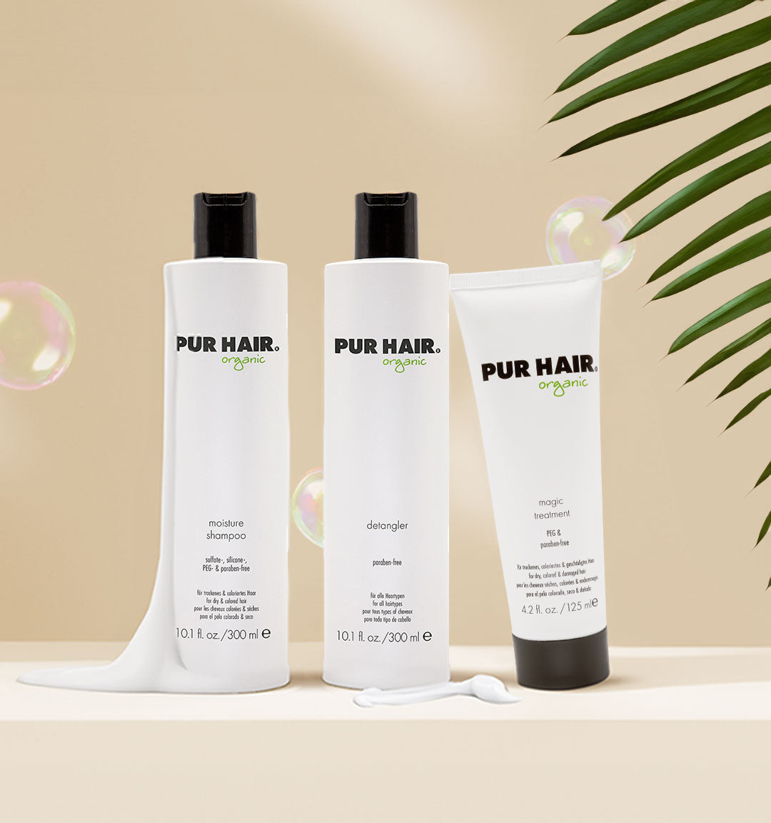 Drei PUR HAIR Organic Produkte – Moisture Shampoo, Detangler und Magic Treatment – auf hellem Hintergrund mit grünen Blättern und Seifenblasen dekoriert