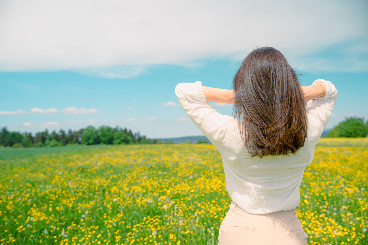 Frau mit glänzendem, glattem Haar steht auf einer Blumenwiese im Sonnenschein – Symbol für gesundes Haar mit natürlichem Glanz