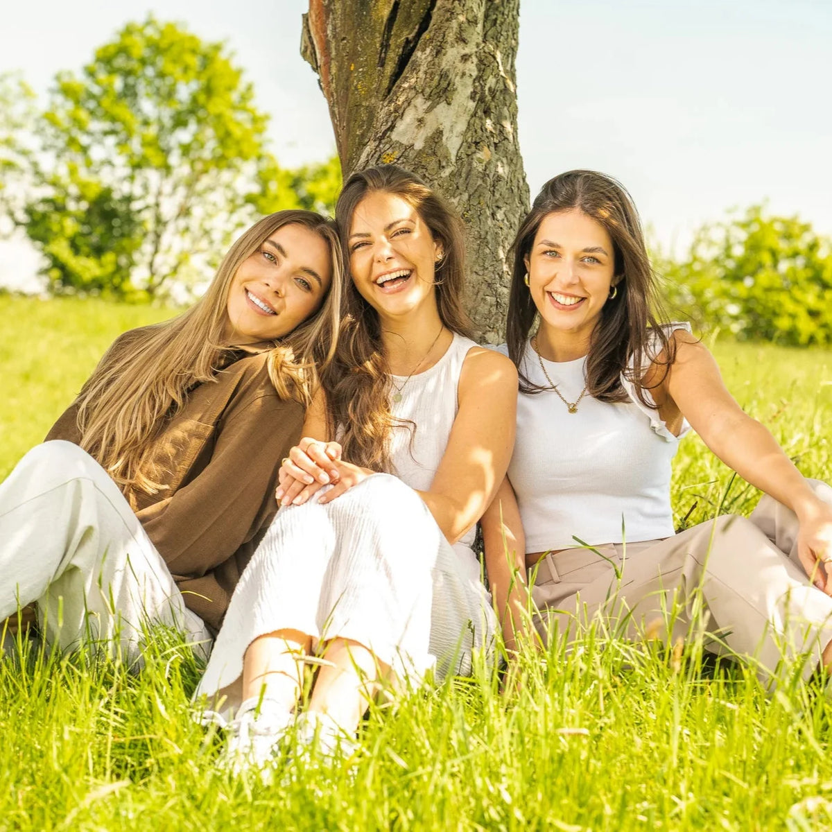 3 lachende Frauen mit heller Kleidung, sitzen an einem Baum auf einer grünen Wiese
