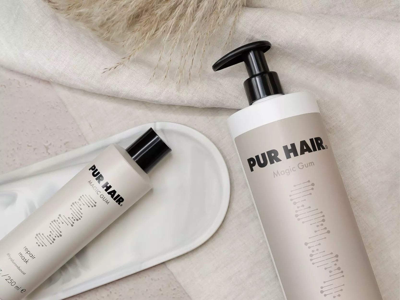 PUR HAIR Magic Gum Repair Serie bestehend aus Shampoo, Repair Mask, dekorativ arrangiert auf grauem Untergrund mit Stoff und Stein-Elementen