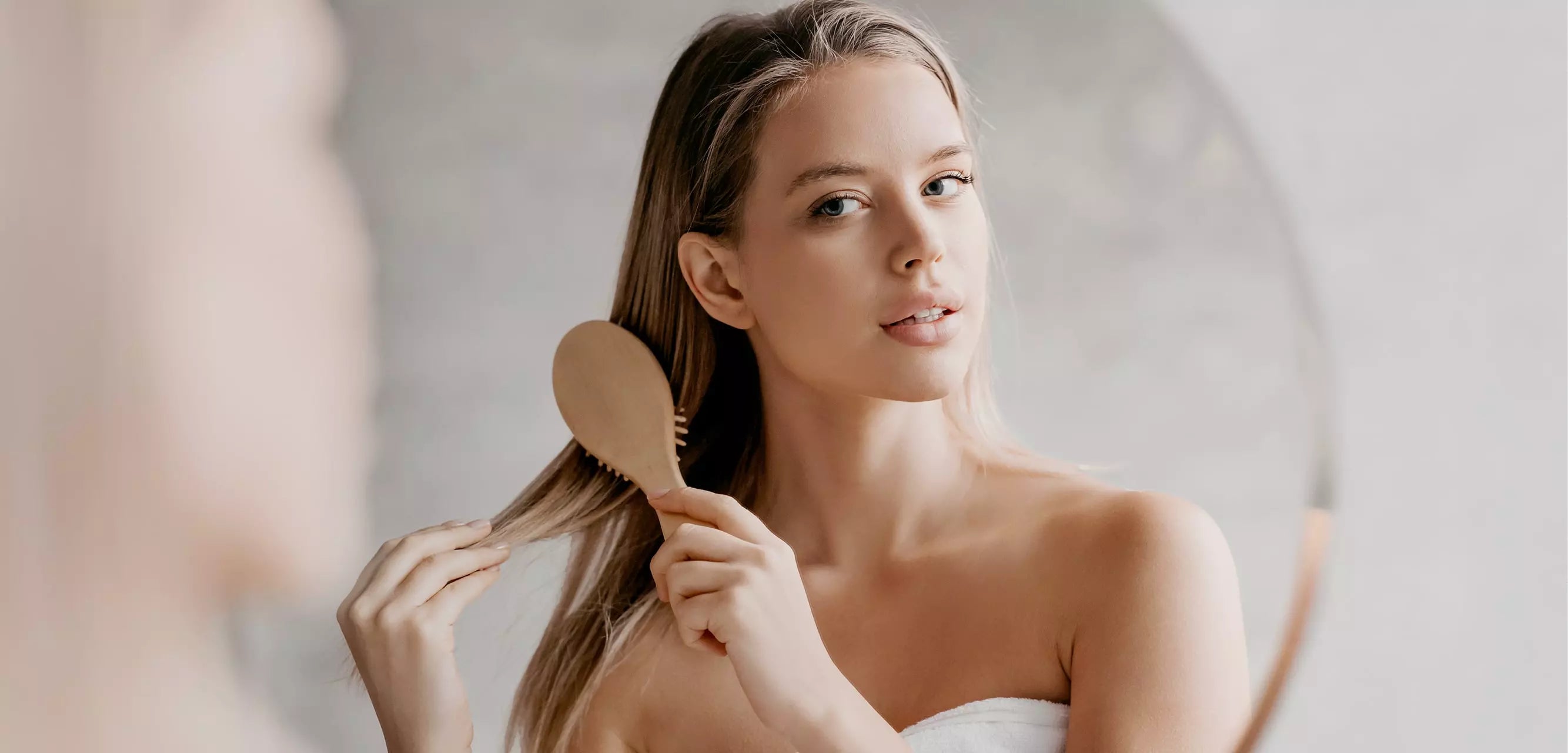 Frau mit langen, blonden Haaren kämmt sich vor dem Spiegel – Haarpflege und tägliche Haar-Routine für gesundes, glänzendes Haar
