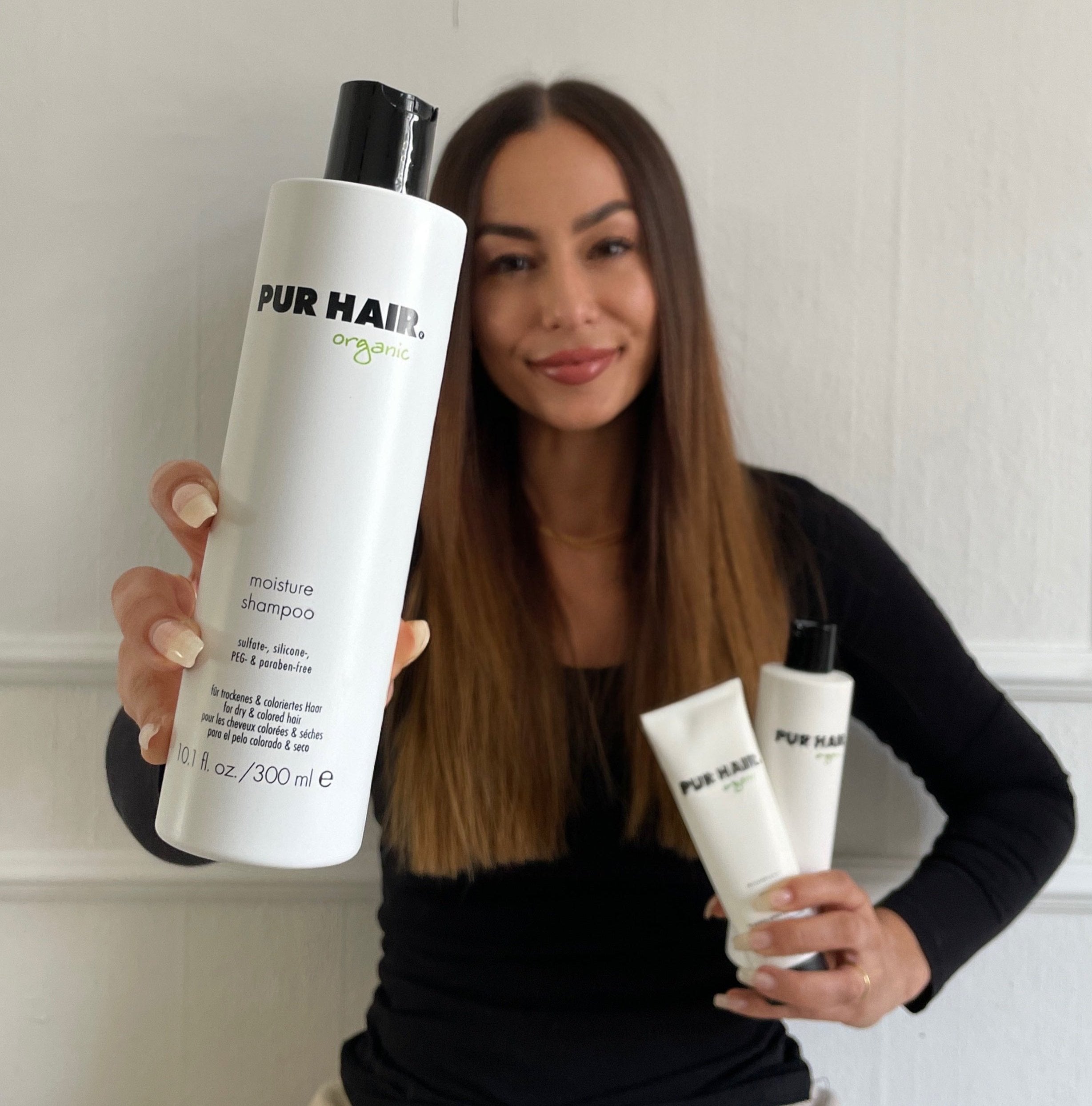 Frau mit langen glatten Haaren hält eine Flasche PUR HAIR Organic Moisture Shampoo in die Kamera, weitere Produkte derselben Serie in der anderen Hand