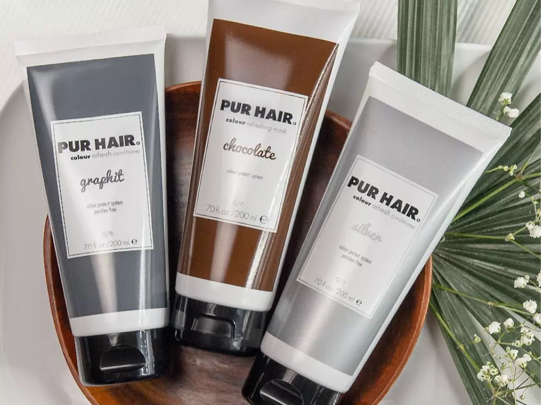 PUR HAIR Colour Refresh Conditioner in den Varianten Graphit, Chocolate und Silber, dekorativ arrangiert in einer Holzschale mit grünen Blättern und weißen Blüten