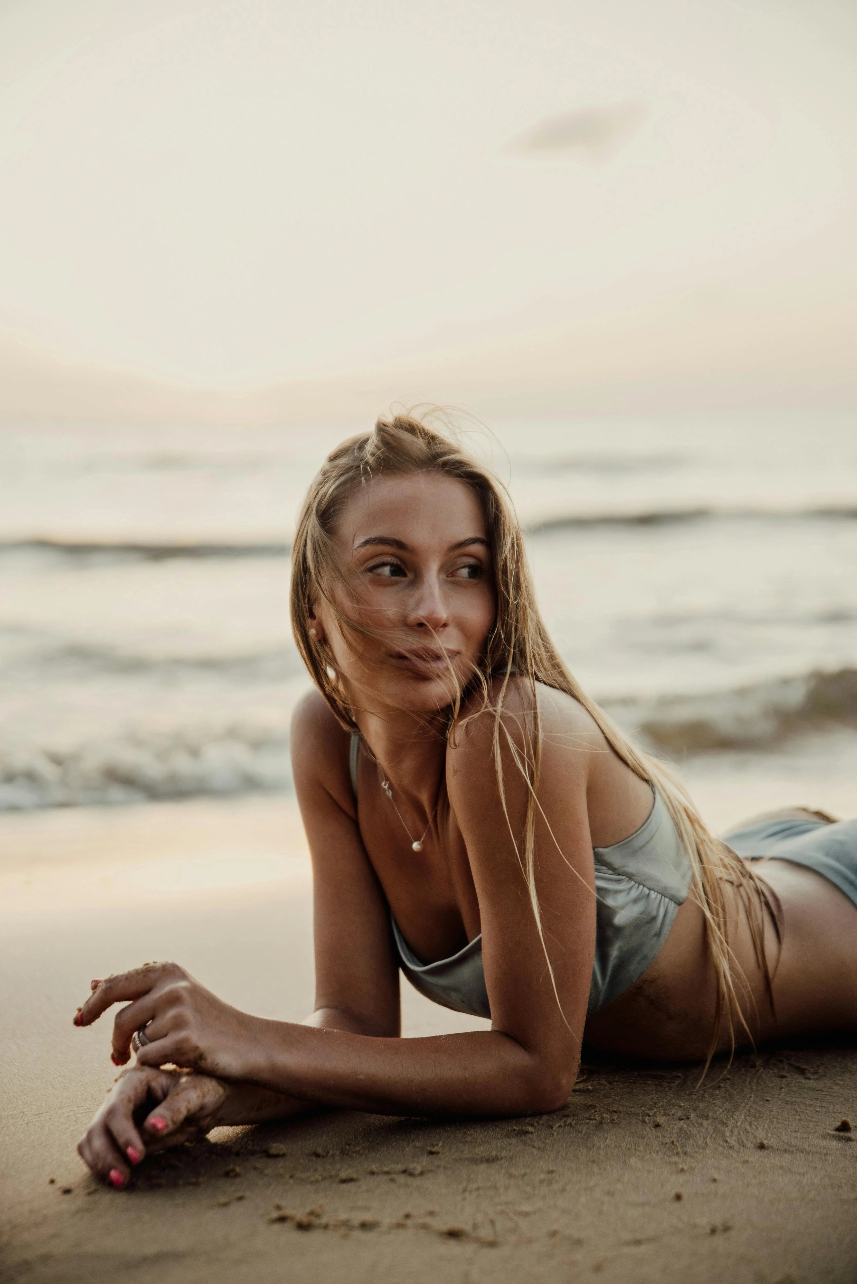 Frau mit langen blonden Haaren, lächelt und schaut zu Seite, liegt am Strand