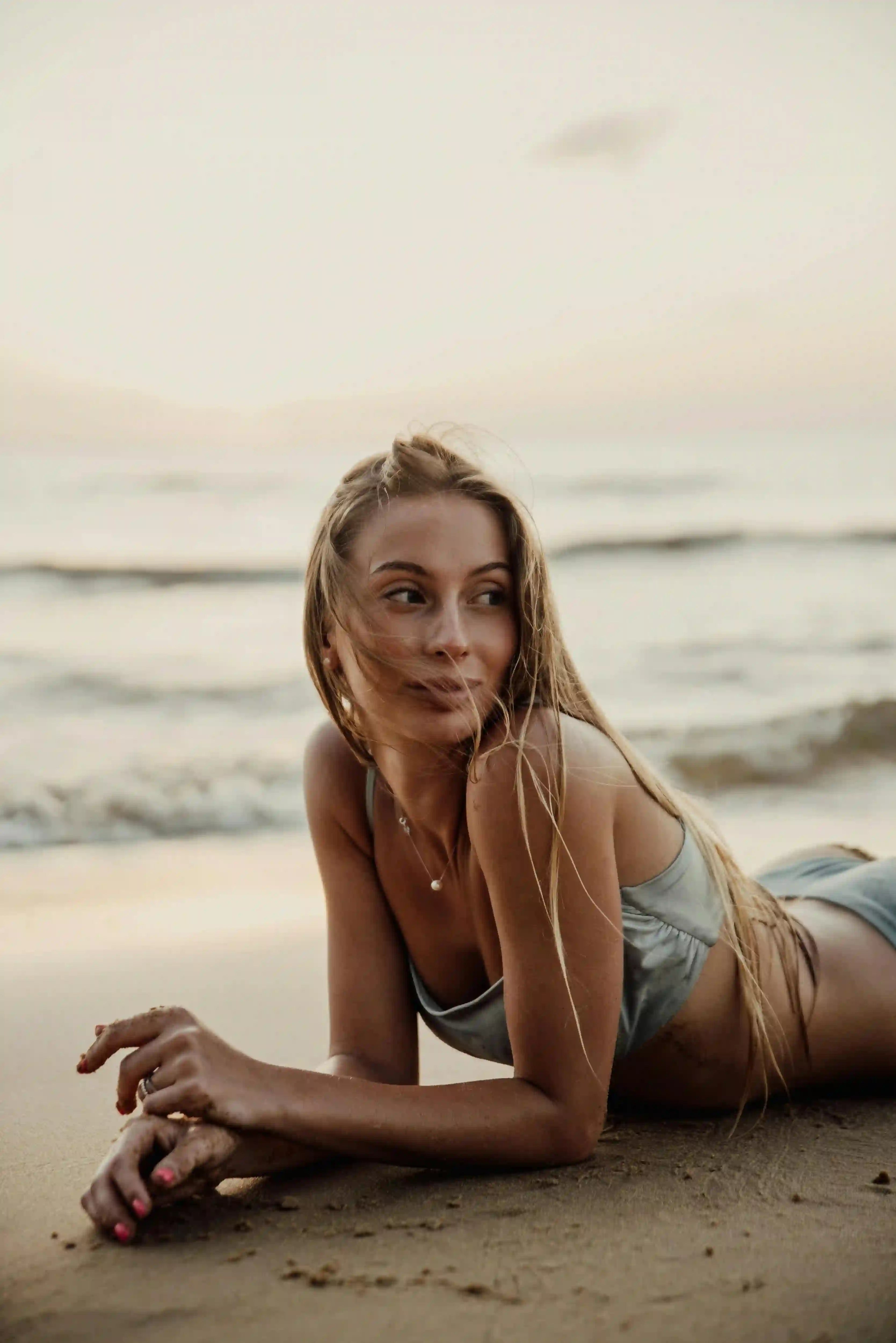 Frau mit langen blonden Haaren, lächelt und schaut zu Seite, liegt am Strand