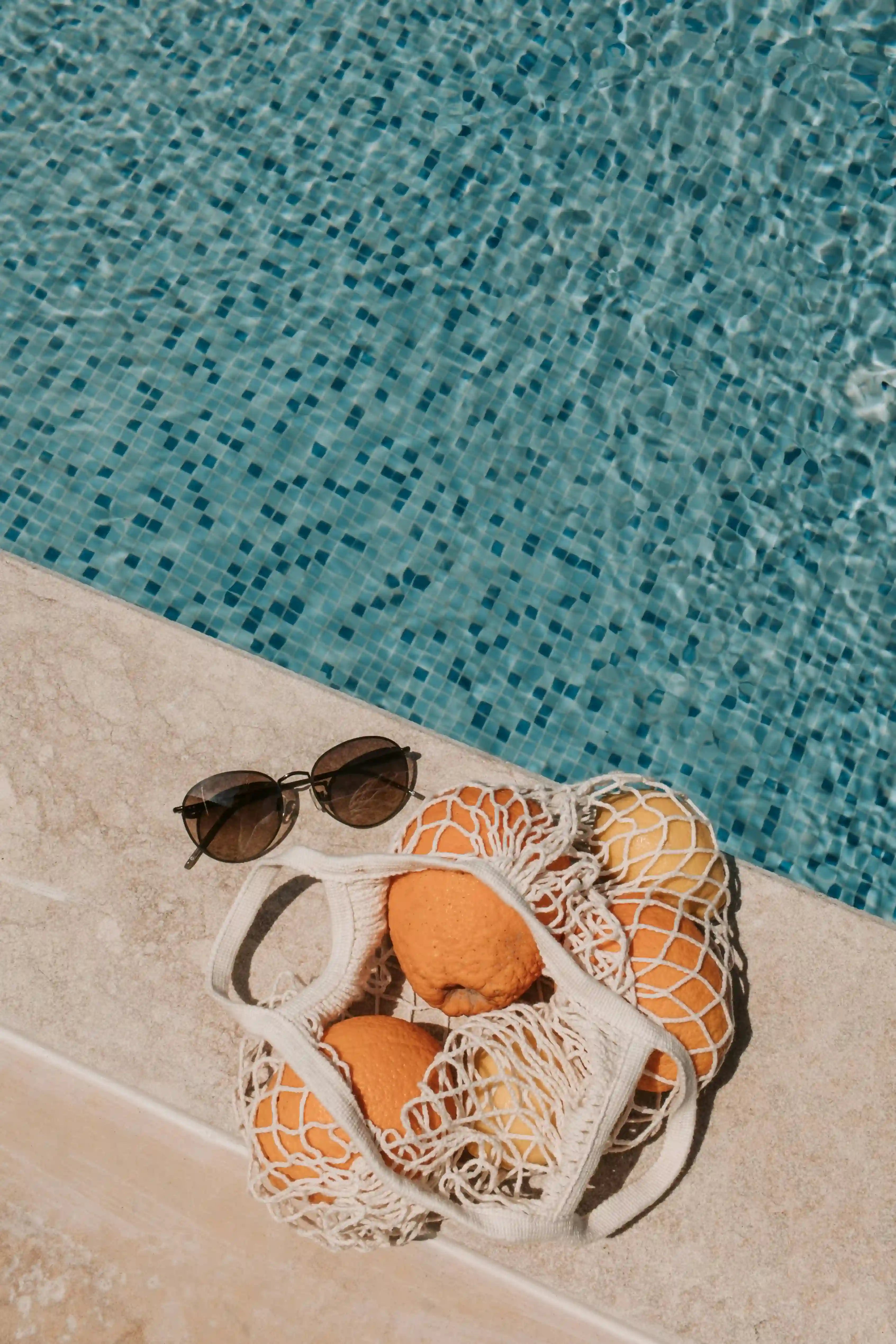 Organgen im Einkaufsnetz, mit Sonnenbrille, liegen neben einem türkisen Pool