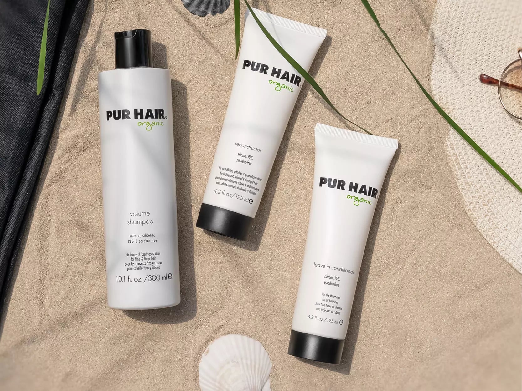 PUR HAIR Organic Haarpflege-Produkte – Volume Shampoo, Reconstructor und Leave-In Conditioner – liegen sommerlich inszeniert im Sand neben Strohhut, Sonnenbrille und Muscheln