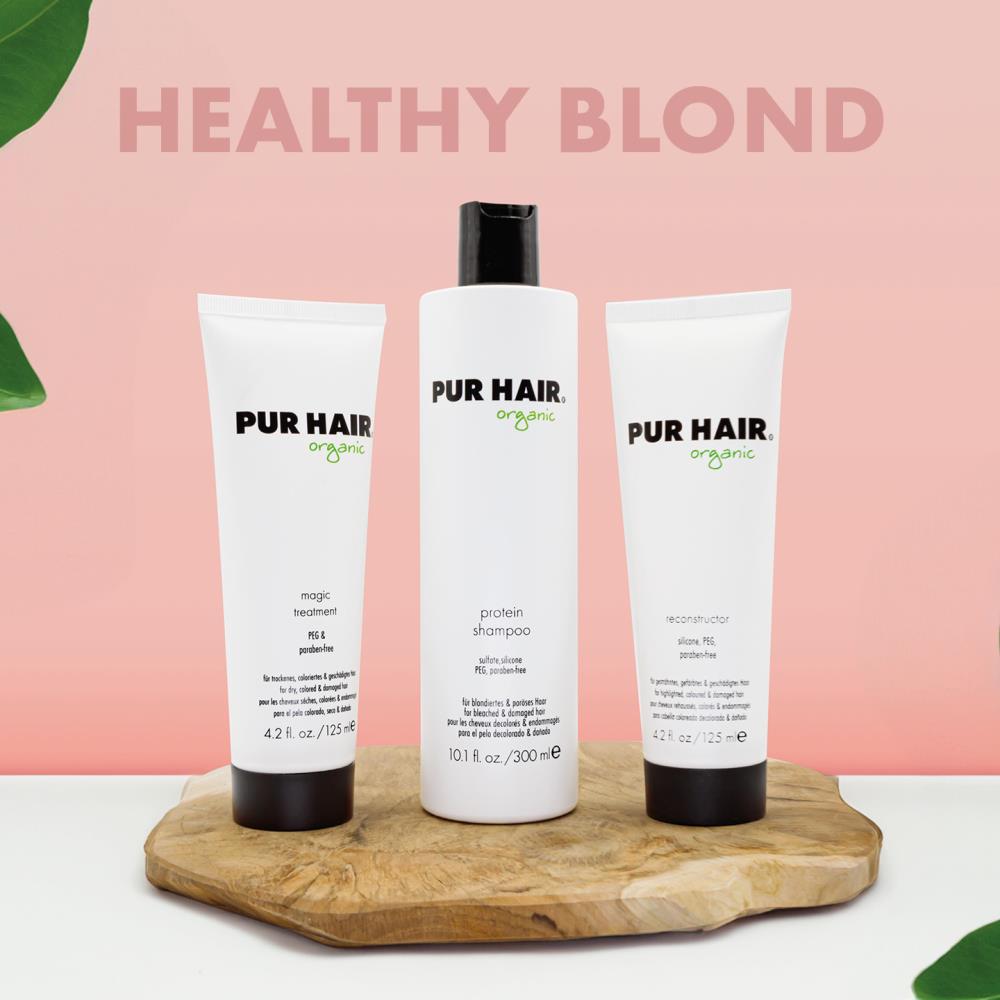 PUR HAIR Organic Blond Set bestehend aus Protein Shampoo, Reconstructor und Magic Treatment. Die drei weißen Flaschen und Tuben stehen dekorativ auf einem Holzbrett vor einem pastell-rosa Hintergrund mit der Aufschrift ‚Healthy Blond‘