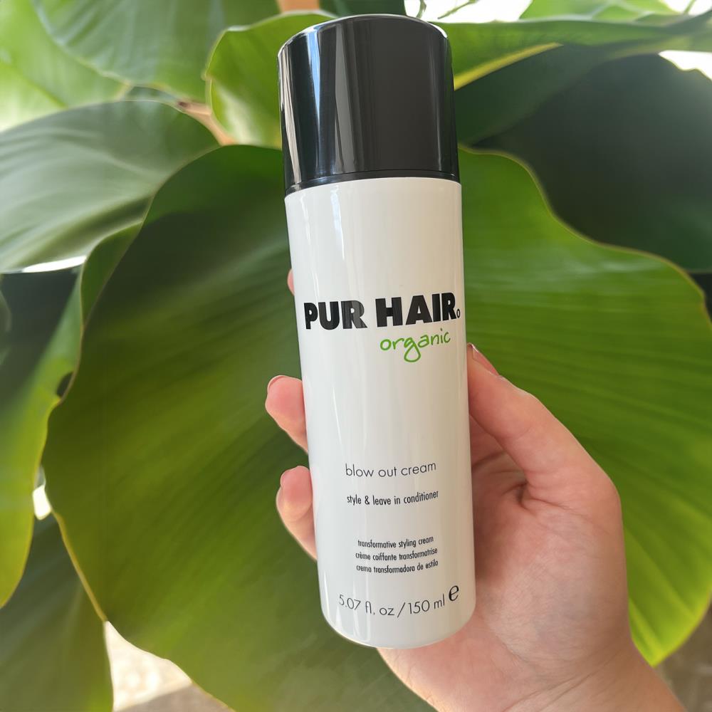 Hand hält PUR HAIR organic Blow Out Cream (150 ml) vor großen grünen Blättern – Leave-in Conditioner und Styling-Creme für glattes, geschmeidiges Haar