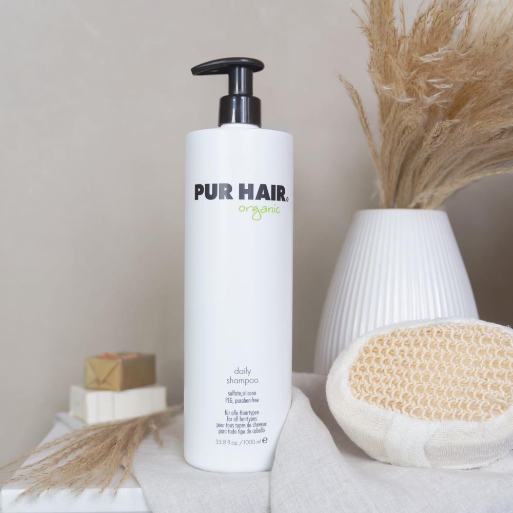 Weiße Flasche PUR HAIR Organic Daily Shampoo, 1000 ml, auf grauem Tuch mit Trockenblumen im Badezimmer platziert
