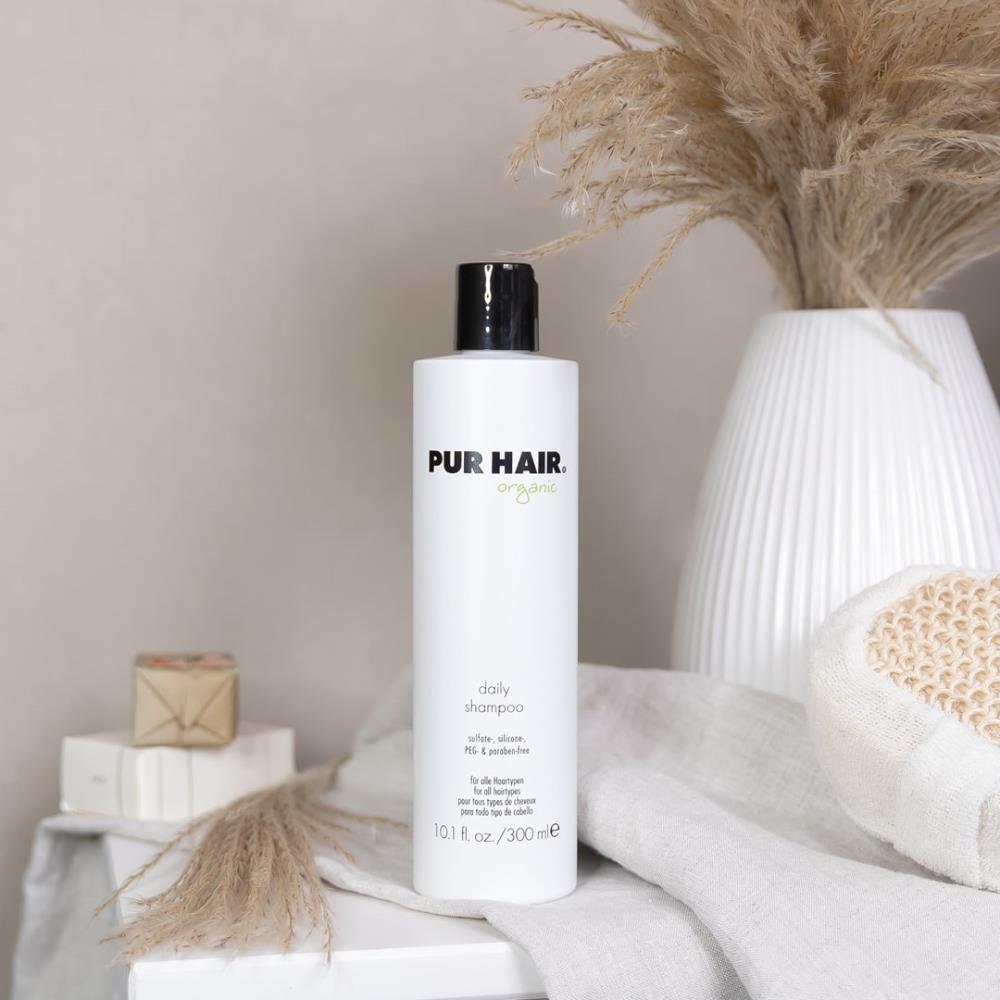 Weiße Flasche PUR HAIR Organic Daily Shampoo, 300 ml, auf grauem Tuch mit Trockenblumen im Badezimmer platziert