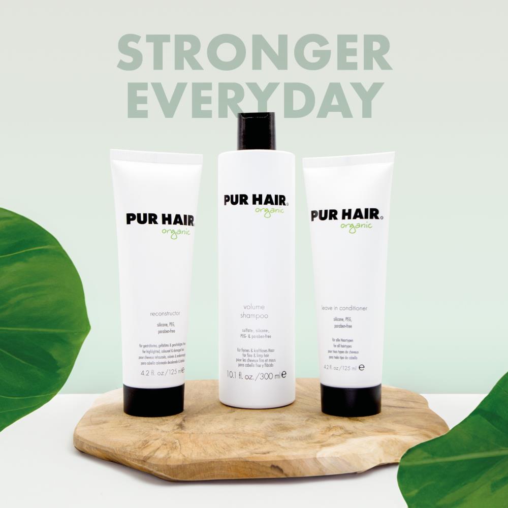 PUR HAIR Organic Feine Haare Set bestehend aus Volume Shampoo, Reconstructor und Leave In Conditioner. Die drei weißen Flaschen und Tuben stehen dekorativ auf einem Holzbrett vor einem pastell-grünem Hintergrund mit der Aufschrift ‚Strong Everyday‘