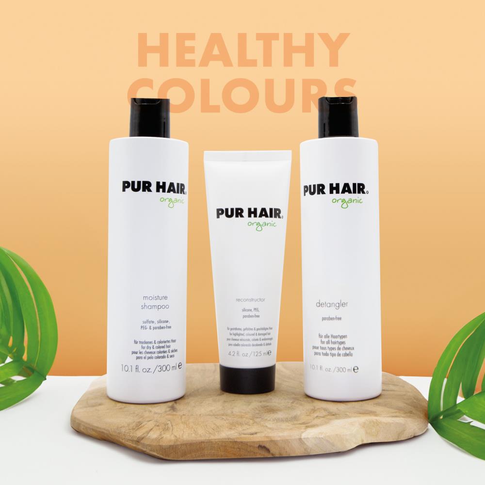 PUR HAIR Organic Colour Protect Set bestehend aus Moisture Shampoo, Reconstructor und Detangler. Die drei weißen Flaschen und Tuben stehen dekorativ auf einem Holzbrett vor einem pastell-orange Hintergrund mit der Aufschrift ‚Healthy Colours‘