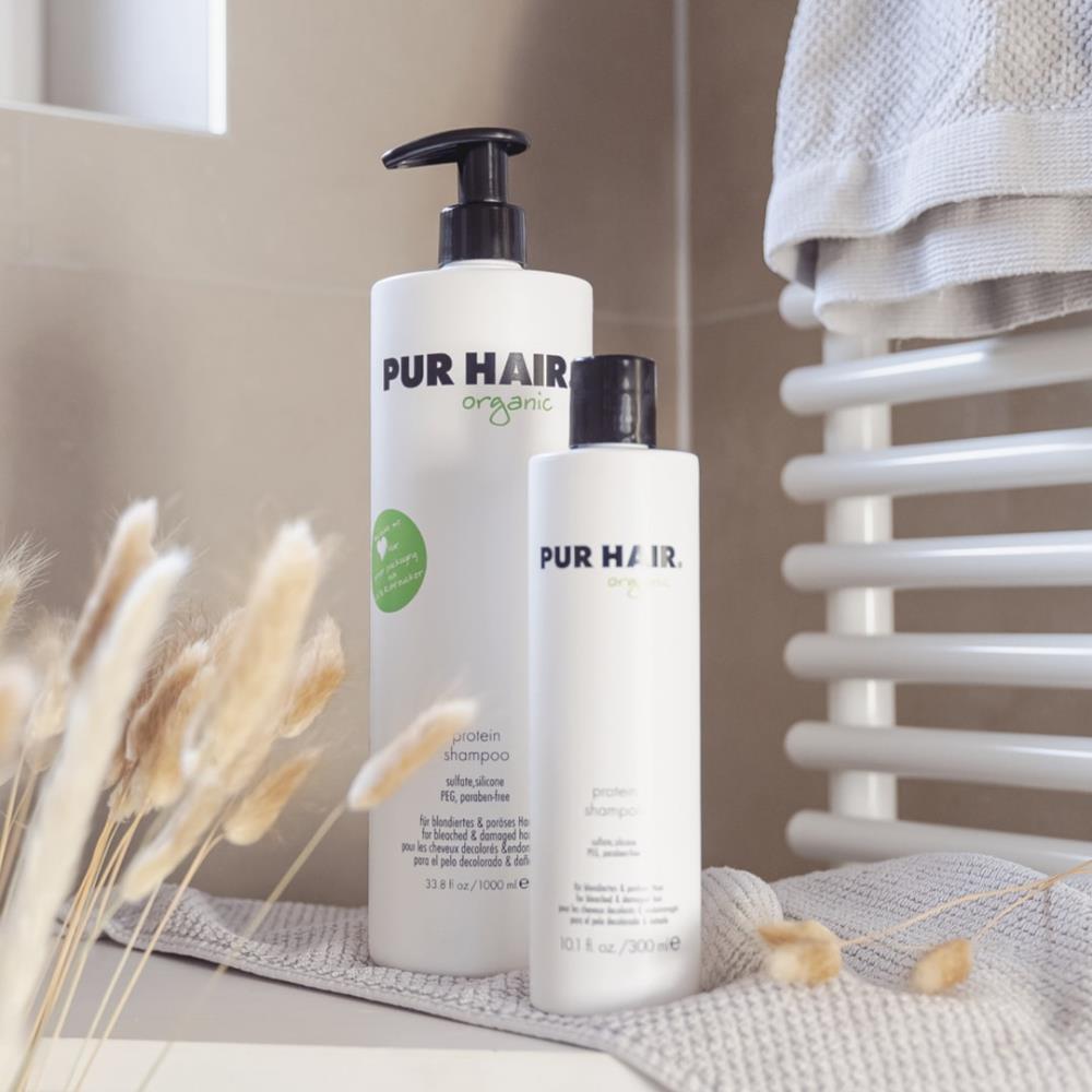 Weiße Flaschen PUR HAIR Organic Magic Treatment 1000 ml und 125 ml, auf grauem Tuch mit Blumen im Badezimmer platziert