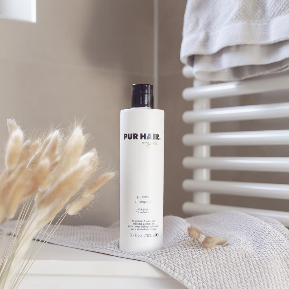 Weiße Flasche PUR HAIR Organic Protein Shampoo, 300 ml, auf grauem Handtuch im Badezimmer platziert