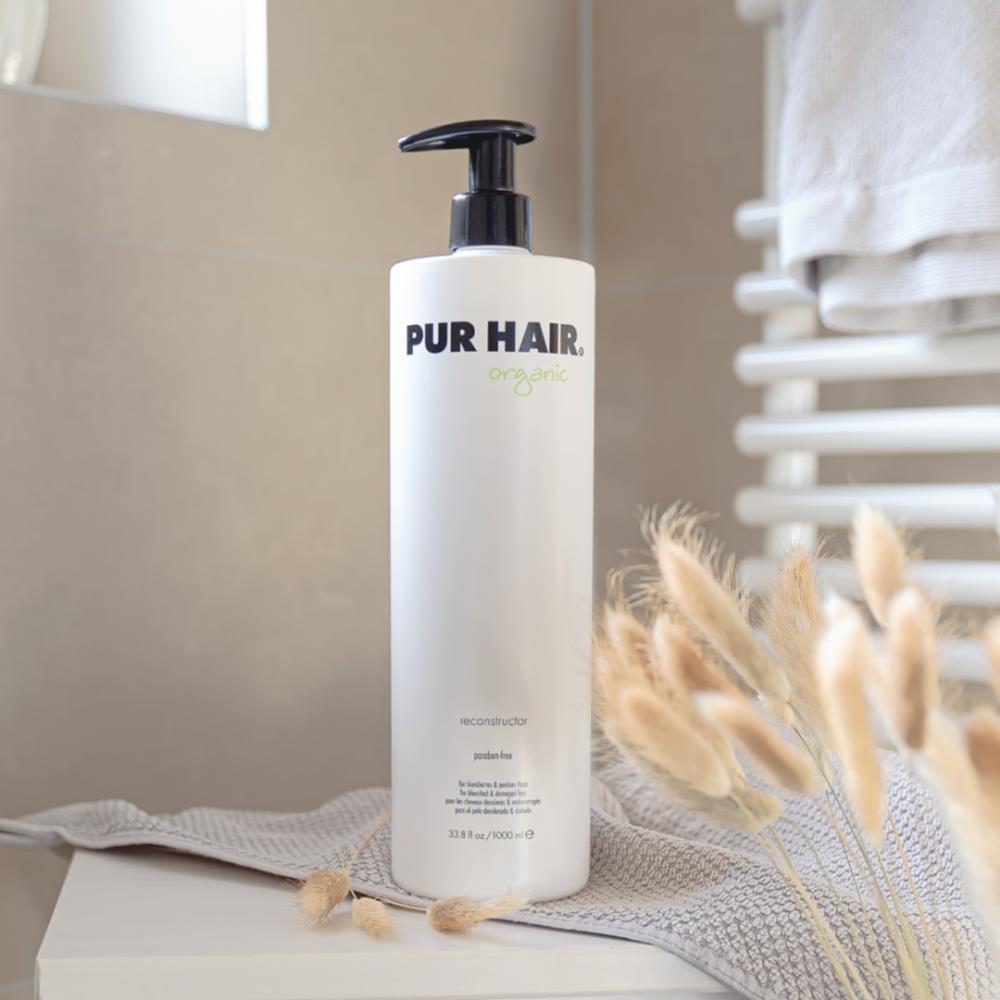 Weiße Flasche PUR HAIR Organic Reconstructor, 1000 ml, auf grauem Handtuch im Badezimmer platziert