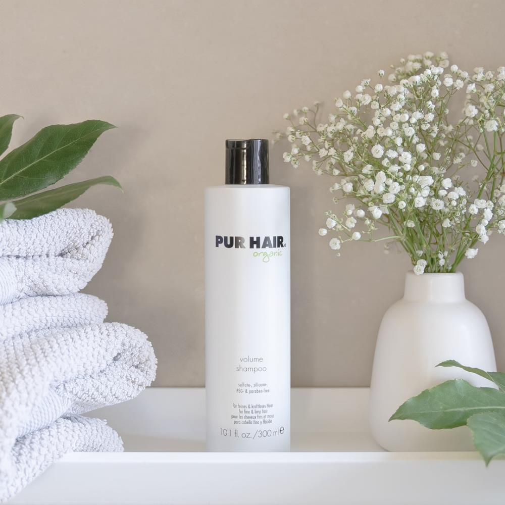 Weiße Flasche PUR HAIR Organic Volume Shampoo, 300 ml, neben grauen Handtüchern mit Trockenblumen im Badezimmer platziert