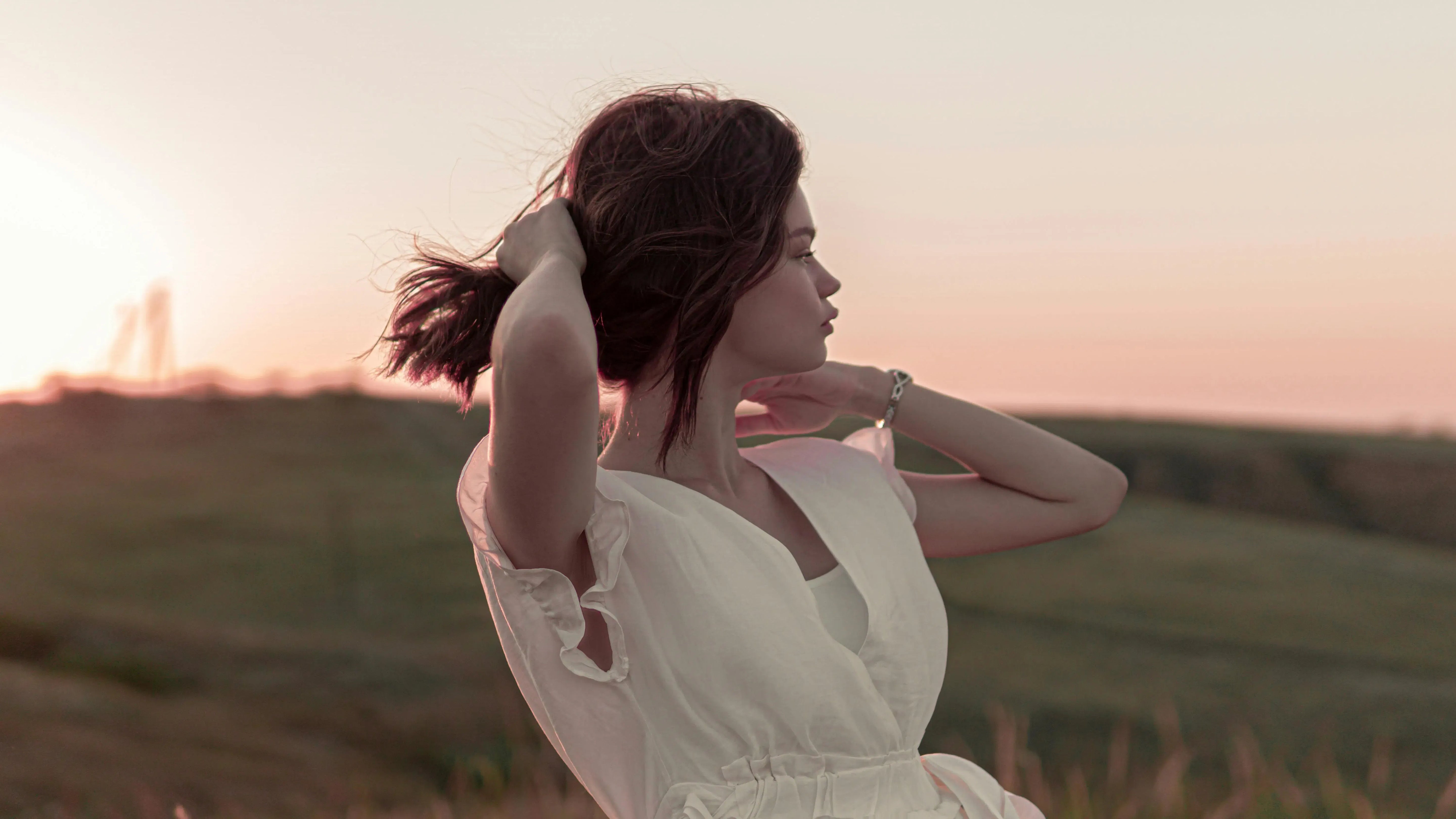 Frau in weißem Kleid, die ihr Haar vor einem Sonnenuntergang über grünen Hügeln hält.
