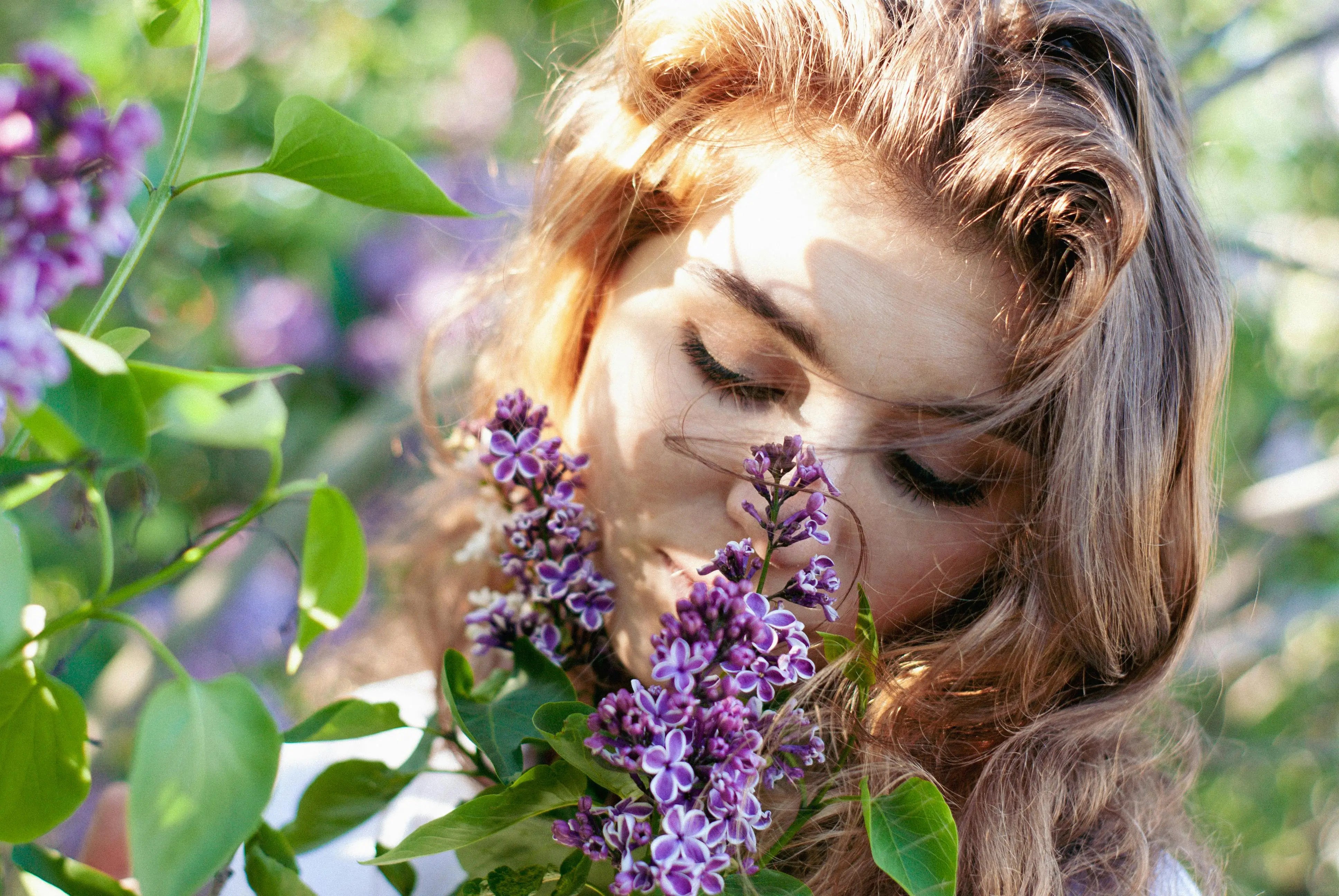 Woman smelling purple lilac flowers in a sunlit garden with green leaves around her.