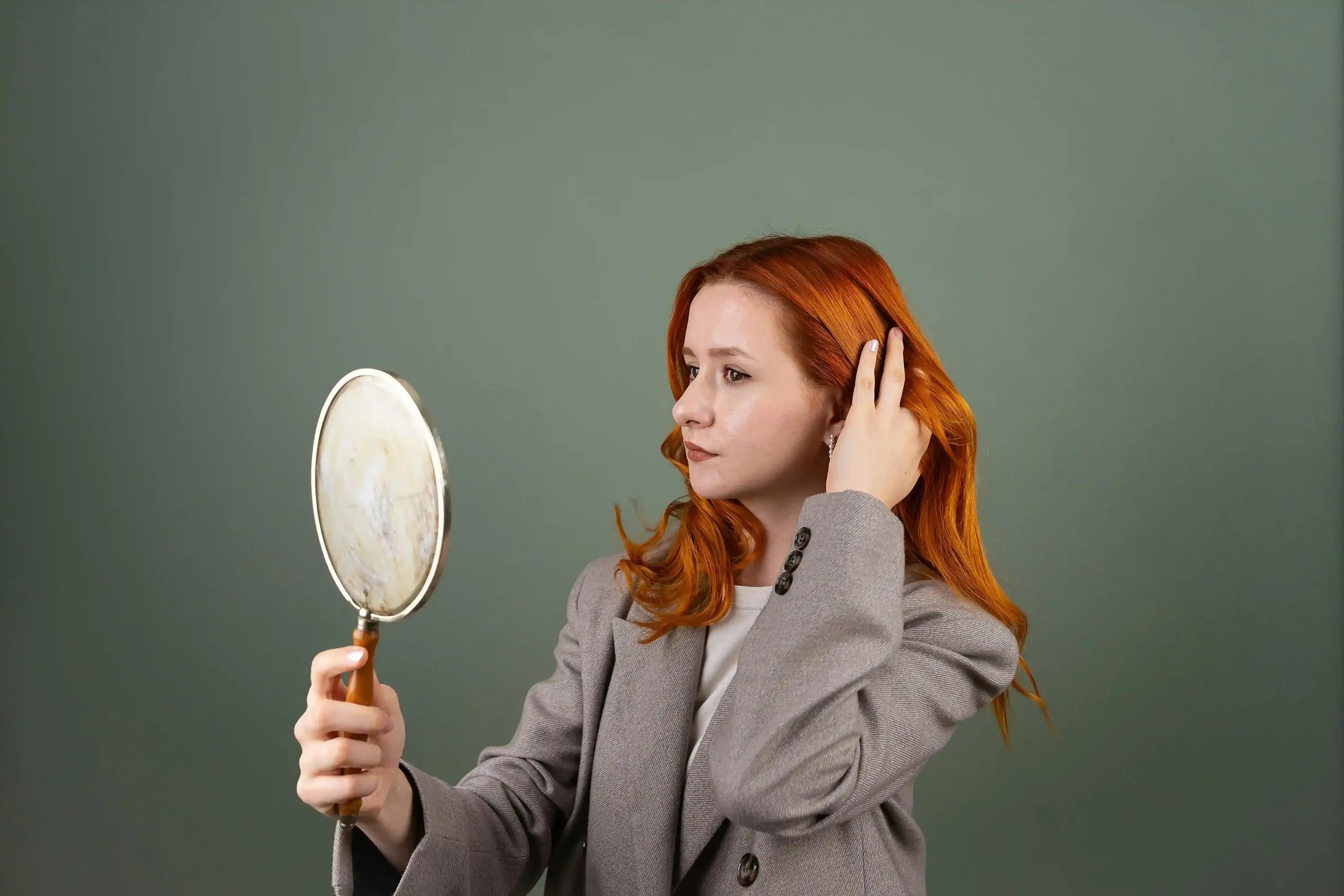 Frau mit roten Haaren, die einen Vintage-Handspiegel hält und ihr Haar vor einem grünen Hintergrund berührt.