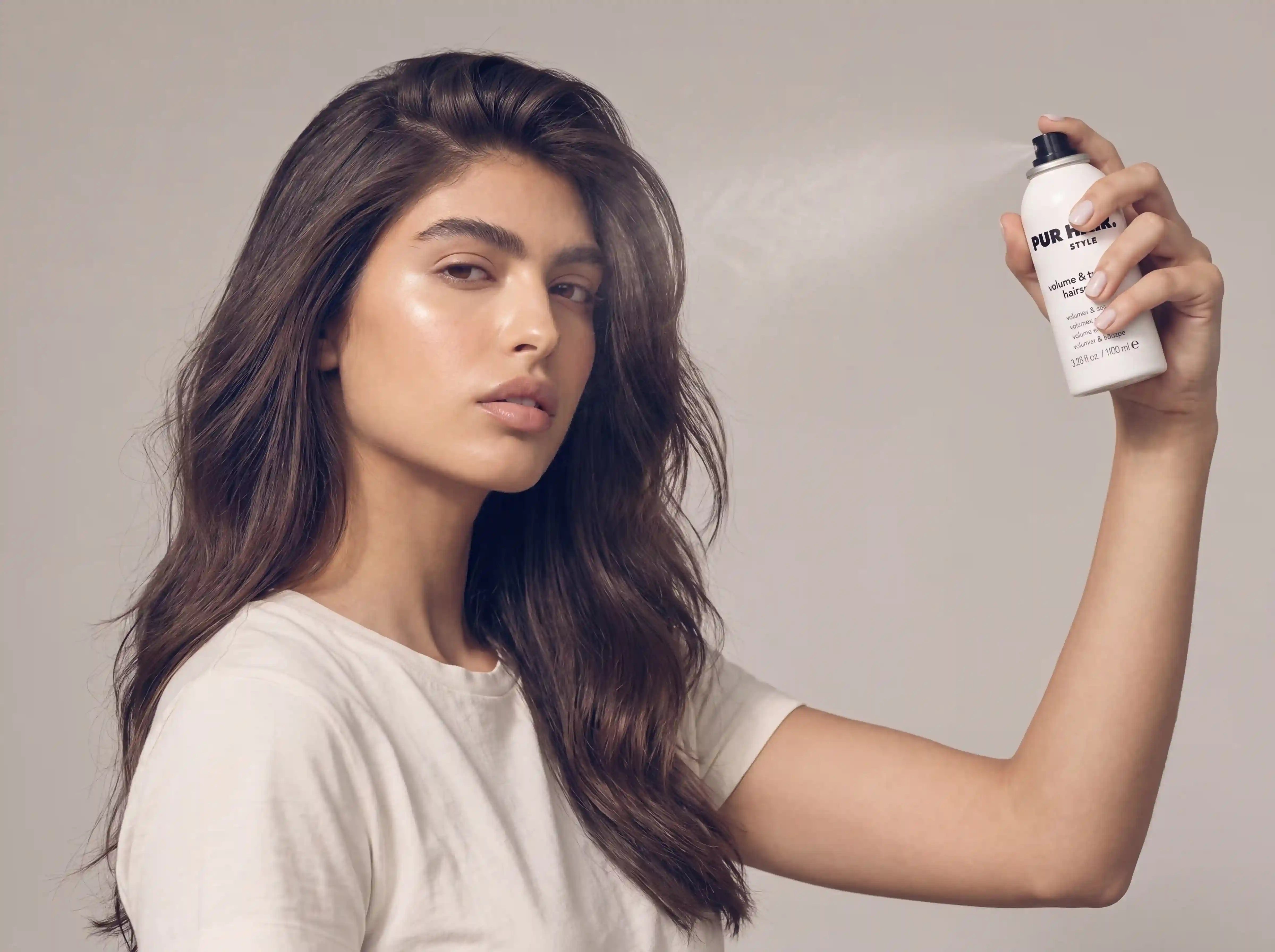 Model mit voluminösem braunem Haar sprüht PUR HAIR Volume & Shine Haarspray für mehr Halt und Glanz auf das Haar.