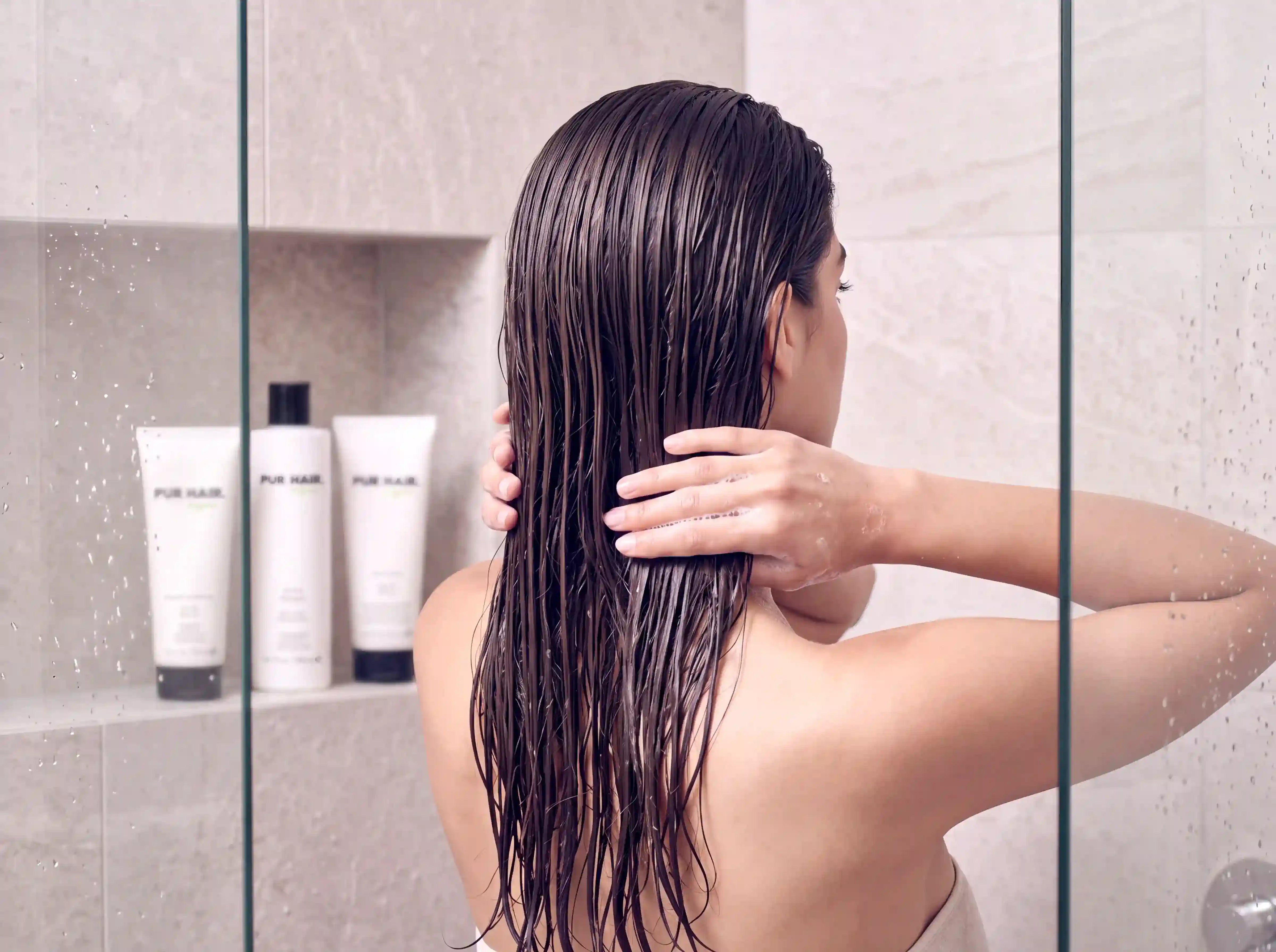 Model arbeitet unter der Dusche eine Haarpflege in das lange nasse braune Haar ein, PUR HAIR Haarpflegeprodukte stehen im Hintergrund.