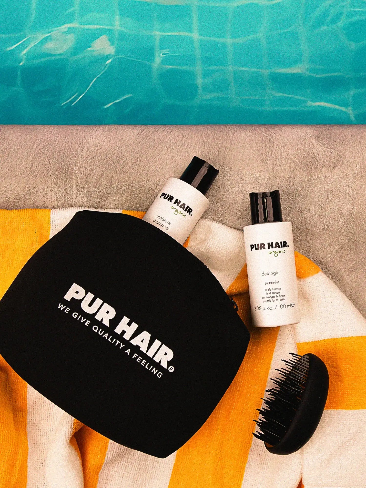 Am Pool liegen auf einem orange-weiß gestreiftem Handtuch, die schwarze PUR HAIR Travel Bag mit der Detangler Brush und Moisture Shampoo 100 ml sowie Detangler 100 ml.