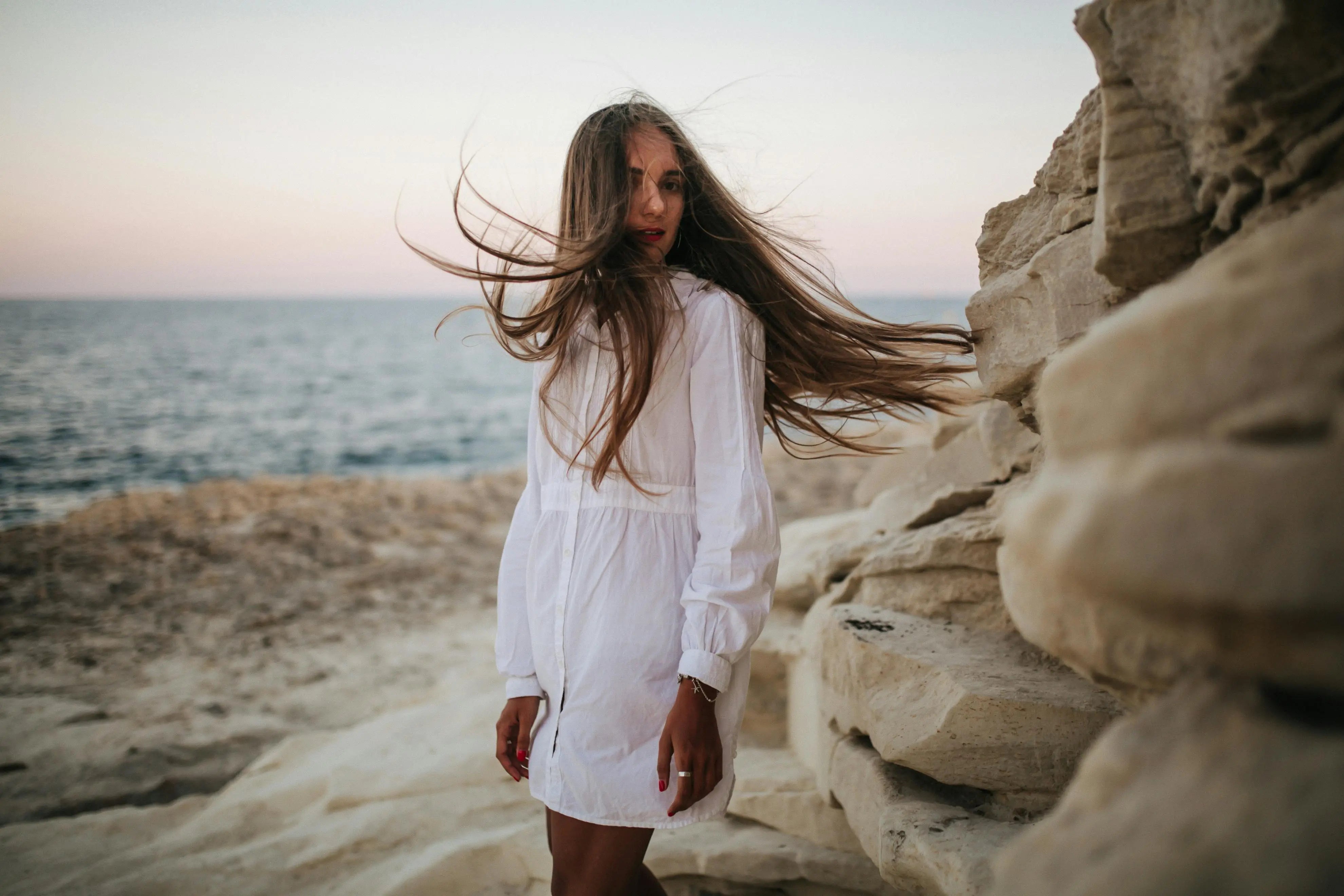 Frau mit langen braunen Haaren, weißem Kleid, Haare wehen im Wind, Frau steht am Meer vor einer Felswand