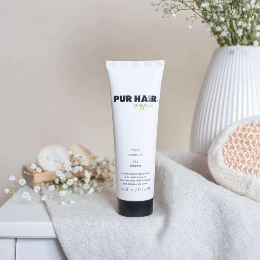 Weiße 125-ml-Tube PUR HAIR Organic Magic Treatment mit schwarzem Deckel, aufgestellt neben Vase und Pflege-Accessoires